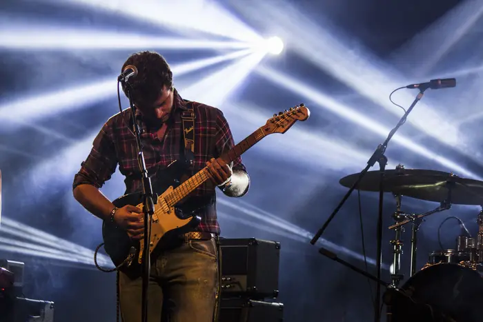Guitarrista de boné em palco, entre feixes de luz branca