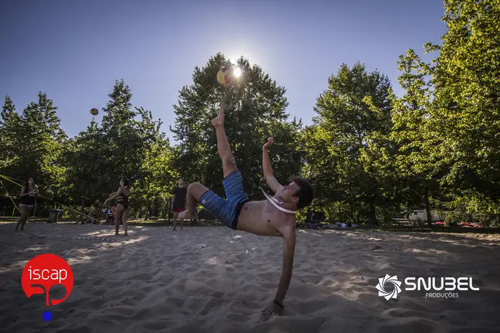 Pessoa num salto acrobático no areal, com árvores ao fundo