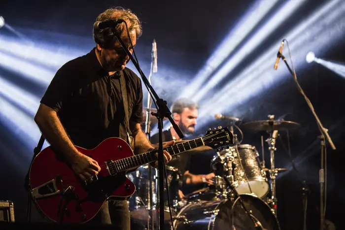 Guitarrista com guitarra vermelha em palco, entre feixes de luz