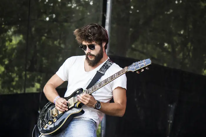 Guitarrista de óculos escuros a tocar ao ar livre, com árvores ao fundo